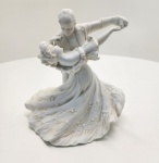 Elegante escultura representando um casal em movimento de dança clássica, modelado em porcelana com acabamento fosco e delicado. A composição captura o instante de uma valsa, com o cavalheiro inclinando suavemente a dama, cuja saia esvoaçante apresenta ornamentos florais em relevo. Os detalhes do traje, gestos e fluidez do movimento conferem à peça uma estética romântica e atemporal, típica da arte decorativa europeia. Dimensões:23 x23 x14 centímetros.