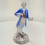 Figura masculina em porcelana Vista Alegre, Portugal, meados do século XX. peça representando cavalheiro do século XVIII, em porcelana moldada e pintada à mão, com trajo típico da época casaca azul cobalto, colete dourado com flores e calças listradas em azul e branco. Base rococó ornamentada com flores em relevo e detalhes dourados. Marca sob a base: V.A. Portugal (Vista Alegre).Dimensões: 26 x 9 x 12 centímetros. Estado de conservação: apresenta reparo visível no pescoço, ausência parcial de flores na base e leve desgaste do dourado.