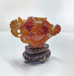 Incensário chinês tripé, inteiramente esculpido em ágata natural, com base original em madeira entalhada. China, c. 19501980.Pequeno vaso trípode de excelente qualidade, talhado em ágata verdadeira de tom âmbar translúcido, com estrias internas e variações naturais típicas da carnelian. O material apresenta brilho vítreo e forte translucidez quando exposto à luz, confirmando a autenticidade da pedra e distinguindo-a do jade e de resinas. A peça exibe alças laterais em forma de dragões ou qilin, habilmente vazadas, e superfície decorada em baixo-relevo. A tampa segue o mesmo padrão de acabamento, e o conjunto repousa sobre base chinesa tradicional em madeira escura, delicadamente entalhada, perfeitamente ajustada ao formato do incensário. Trabalho de notável refinamento dentro da produção chinesa em hardstone da segunda metade do século XX, com boa simetria, polimento fino e excelente presença visual. Dimensões: 11x11x7 centímetros. Apresenta pequenos desgastes e perdas em pontas mais delicadas, comuns em esculturas de pedra. dimensões: 11x11x7 centímetros.