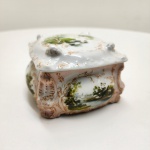 Caixa de formato quadrangular com tampa ajustada, em porcelana moldada e esmaltada, decorada com pinturas manuais de paisagens campestres nas laterais e na tampa, envoltas por arabescos dourados em relevo. Apresenta bordas sinuosas e decoração rocaille, típica do gosto Rococó que marcou a produção de porcelanas alemãs e austríacas no final do século XIX. Interior e base esmaltados em branco. Peça de fino trabalho artesanal, representativa da tradição decorativa da Europa Central. Dimensões: 9 x 13 x 13 centímetros.