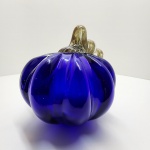 Elegante escultura em vidro artístico soprado, em formato de abóbora estilizada, de corpo facetado e tom azul cobalto profundo, com aplicação superior em vidro retorcido de tonalidade azul escuro translúcido. A peça revela domínio técnico excepcional na manipulação do vidro ainda em fusão, evidenciado na simetria das gomos, no brilho intenso e na pureza do vidro. O contraste entre o corpo liso e a alça retorcida cria um efeito visual vibrante, típico das criações modernas inspiradas na escola Murano, onde o vidro é simultaneamente matéria escultórica e luminosa. O azul cobalto uma das cores mais difíceis de obter com transparência e uniformidade é aqui explorado em toda sua profundidade, resultando em uma obra de grande presença visual e sofisticação cromática. Altura: 17 x 17 centímetros. Estado de conservação: Apresenta pequena perda na ramagem.