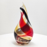 Vaso escultórico em murano artístico soprado, de forma orgânica e torcionada, apresentando composição cromática em vermelho rubi, âmbar e preto fumê, entrelaçados em faixas espiraladas que se fundem no corpo translúcido de base incolor. A obra revela a técnica de sommerso (vidro sobreposto), característica das oficinas de Murano, onde camadas de vidro colorido são encapsuladas em cristal límpido, produzindo efeitos de profundidade e movimento interno. A espessura generosa do vidro e o brilho impecável do acabamento indicam execução manual de alto nível, com polimento em fogo e modelagem livre, sem o uso de moldes. A silhueta alongada e o ápice levemente curvado conferem à peça uma presença escultórica e dinâmica, típica da produção artística veneziana das décadas de 1950-1970, quando o design em vidro alcançou pleno reconhecimento internacional.Dimensões:42 x 20 x 12 centímetros.