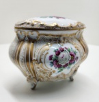 Caixa em porcelana Rosenthal / Bavaria, Alemanha. Elegante caixa com tampa em porcelana fina alemã, de corpo bojudo e bordas recortadas, apoiada sobre quatro pés curvos. Decoração em flores policromadas rosas e anêmonas sobre fundo branco, com detalhes em ouro pintado à mão e finos arabescos dourados em pontilhismo. Tampa com composição floral central e frisos em relevo com douração intensa. Marca em verde sob a base: Rosenthal / Bavaria, com inscrição manuscrita, correspondente à linha decorativa. Dimensões:15x18x13 centímetros.