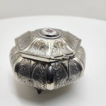 Elegante caixa de formato octogonal executada em metal prateado, repousando sobre pés de apoio ornamentados. A superfície apresenta ricamente gravados motivos fitomórficos em relevo, com destaque para folhas estilizadas em padrão simétrico, típicos do estilo neoclássico tardio / art nouveau de influência europeia. Na tampa, medalhão central oval em porcelana esmaltada com delicada pintura floral em tons de marrom, amarelo e branco, realçando o caráter artesanal da peça. Interior liso, com leve pátina do tempo que realça a autenticidade.Dimensões:10 x 16 centímetros.