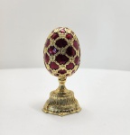 Elegante ovo decorativo no estilo Fabergé, adquirido na Rússia e elaborado em liga metálica de qualidade com banho de ouro. Seu exterior é revestido com esmalte translúcido em tom rubi imperial, aplicado sobre base texturizada em padrão guilloché, criando profundidade e reflexos típicos das peças inspiradas na joalheria imperial russa. A superfície exibe arabescos e elementos folheados em ouro, formando uma treliça rica em detalhes. A abertura articulada revela interior esmaltado em rubi profundo. A peça apresenta duas surpresas, seguindo a tradição Fabergé. Dimensões: 12x6 centímetros.