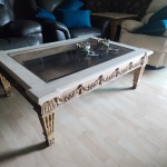 Mesa de centro executada em madeira entalhada, decorada com frisos, folhas de acanto e guirlandas florais em relevo, em clara referência ao estilo Louis XVI. A peça apresenta pernas estriadas com base ornada e entalhes profundos de excelente presença visual. O tampo é composto por placa de mármore claro com borda chanfrada, ladeando centro em vidro plano embutido, solução que preserva a visão da estrutura entalhada inferior. Acabamento com pátina envelhecida original, apresentando desgaste natural do tempo que confere caráter autêntico à peça.