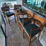 Conjunto composto por mesa redonda em madeira maciça, com tampo formado por ripas em composição geométrica, apoiado sobre base cruzada. Acompanha quatro banquetas altas, cada uma com braços e encosto vazado, decorado com entalhe balinês em forma de raios solares, motivo típico da iconografia artesanal de Bali. Os pés apresentam torneamento delicado, e os assentos são estofados em tecido verde mesclado, conferindo conforto e harmonia ao conjunto. Dimensões: mesa 1,00x0,80., cadeiras 1,16x0,55x0,51.