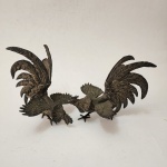 Par de esculturas decorativas representando galos em combate, confeccionadas em metal banhado a prata, com detalhamento minucioso de plumagens, asas abertas e caudas elevadas que conferem grande dinamismo e teatralidade à composição. O acabamento em prata realça os volumes e dá luminosidade à modelagem, mantendo pátina elegante. Conjunto típico da produção decorativa oriental do século XX. Dimensões: 20x22x18 centímetros. ( cada)