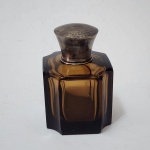 Elegante perfumeiro Art Déco, cerca 19201940, modelado em cristal fumê espesso e finamente lapidado em linhas geométricas, com cantos recortados e facetas típicas da estética modernista do período. A peça é coroada por tampa em prata de lei, trabalhada com delicado acabamento martelado, revelando refinamento técnico e equilíbrio entre materiais. Perfumeiro de toucador europeu, de presença discreta e sofisticada, representativo da produção de luxo das primeiras décadas do século XX. Dimensões: 11x7 centímetros.