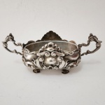 Linda terrina decorativa em metal banhado a prata, exuberantemente trabalhada no estilo rococó, com profusa ornamentação em relevo composta por volutas, cartelas e elementos assimétricos de forte dinamismo. Corpo oval apoiado sobre quatro pés modelados, e ladeado por duas asas vazadas de desenho sinuoso, conferindo elegância e movimento ao conjunto. Interior liso e borda recortada, demonstrando refinamento técnico e decorativo. Peça de presença marcante, representativa da ourivesaria decorativa europeia do final do século XIX ao início do XX.
