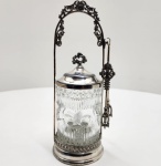 Pickle Castor em vidro moldado e metal prateado Europa, fim do século XIX. Charmoso pickle castor (compoteira com suporte) composto por recipiente em vidro moldado transparente, com decoração em relevo de faixas verticais e reservas florais, acompanhado de tampa e estrutura em metal prateado finamente trabalhada com motivos florais e rocalhas. O conjunto é sustentado por arco decorativo em estilo vitoriano, com ornamentação vegetalista e suporte lateral para pinça metálica, de fino acabamento. A peça reflete o gosto e a sofisticação das criações domésticas europeias do final do século XIX, quando a mesa passou a incorporar objetos artísticos de uso cotidiano, aliando praticidade e ornamentação refinada. Dimensões: 26 x 9 centímetros.