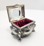 Elegante caixa decorativa em metal prateado, trabalhada em estilo Rococó com folhas, volutas e motivos florais em alto-relevo. O tampo apresenta composição central emoldurada com flores e ornamentos em estilo europeu clássico. As laterais exibem relevos ondulados e pés recurvados, típicos da estética Rocaille. O interior é revestido em veludo vermelho. Dimensões: 5 x 7 x 4 centímetros.