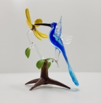Escultura em vidro artístico Murano Beija-flor e flor amarela Delicada escultura em vidro artístico de Murano, representando um beija-flor azul em pleno voo a sorver o néctar de uma flor amarela, sobre base em forma de galho transparente com folhas verdes e tronco em tons âmbar. Modelada e soprada manualmente, a peça revela a transparência e o brilho característicos do vidro veneziano, com perfeita harmonia de cores e movimento dinâmico que transmite leveza e poesia. Altura: 14 x 10 x 7 centímetros.