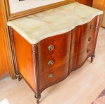 Cômoda inglesa em madeira nobre, de frente sinuosa e acabamento primoroso, apresentando entalhes em folhas estilizadas nas pilastras e filetes de marchetaria acompanhando o desenho das gavetas. O tampo, em alabastro maciço, possui bordas suavemente modeladas e veios naturais em tons claros e dourados. A peça conta com quatro gavetas curvas, todas com puxadores metálicos originais, e pés torneados e estriados com ponteiras metálicas patinadas. Conserva excelente integridade estrutural e acabamento característico do mobiliário inglês da primeira metade do século XX. Dimensões 1,00x1,00x0x0,70.