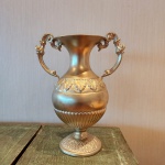 Vaso decorativo em metal com acabamento dourado, apresentando formato clássico com corpo ovóide, bojo ornamentado por friso em relevo de folhas estilizadas e base canelada. Alças laterais simétricas, em volutas decorativas, reforçam a estética tradicional. Peça de presença, com brilho homogêneo e excelente apelo decorativo. Dimensões: 32x26x16 cencentímetros.