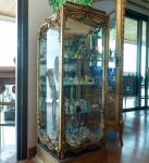 Elegante vitrine em madeira entalhada, executada em estilo Luís XVI, ricamente ornamentada e com estrutura envidraçada em todas as faces. A cinta superior apresenta tampo em mármore branco suavemente patinado, apoiado sobre molduras douradas com frisos em relevo.O corpo da vitrine é decorado com festões florais, guirlandas pendentes, medalhão oval central e amarras clássicas, todos esculpidos em madeira e recobertos por douração com pátina envelhecida. Os cantos recebem folhas, laços e volutas típicas do repertório neoclássico francês do final do século XVIII.O interior possui prateleiras de vidro e fundo espelhado, conferindo amplitude e realce às peças expostas. A base em madeira reforçada mantém o conjunto elevado e estável, preservando a imponência da peça. Dimensões:1,30x0,45x0,45 metros.