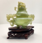 Incensário chinês tripé com tampa, inteiramente esculpido em jade nefrita natural, China, meados do século XX. Peça de formato tradicional, composta por corpo circular apoiado em três pés curvos, alças laterais em forma de dragões e tampa encimada por leão guardião (fu dog). O jade apresenta cor verde uniforme, com translucidez suave e textura compacta características típicas da nefrita verdadeira, pedra venerada na cultura chinesa pela dureza, pureza e significado simbólico. O entalhe revela trabalho artesanal cuidadoso, com curvas limpas, equilíbrio nas proporções e boa definição nas áreas vazadas das alças. A peça repousa sobre base original em madeira escura, delicadamente entalhada, no estilo tradicional chinês. Modelo clássico de censer em jade, produzido na segunda metade do século XX, preservando a estética e o simbolismo das peças antigas utilizadas em templos e residências nobres. Dimensões: 17x15x10 centímetros. Apresenta pequenos desgastes e perdas em pontas mais delicadas, comuns em esculturas de pedra.