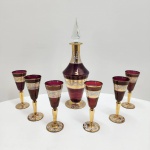 Conjunto composto por licoreira e seis taças em cristal turco, trabalhado em técnica overlay rubi (camada de cristal colorido sobre base transparente) com rica aplicação de ouro e esmaltação decorativa. A licoreira apresenta stopper facetado em cristal transparente e corpo em rubi intenso, ornamentado com faixas largas de ouro polido e arabescos em esmalte branco com detalhes pontilhados.As seis taças seguem o mesmo padrão decorativo, com bojo rubi e hastes completamente revestidas em ouro, além de bases decoradas com motivos em esmalte e ouro, típicos da produção turca