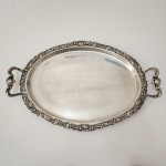 Belo tabuleiro oval em metal prateado, manufatura Meridional E.P.S, apresentando rica borda trabalhada com ornamentação em relevo de inspiração rococó, composta por volutas, concheados e folhagens entrelaçadas. Alças laterais vazadas, de desenho sinuoso e equilibrado, integrando-se harmonicamente ao conjunto. Superfície ampla e lisa, ideal para serviço ou composição decorativa. Peça de elegante presença e excelente estado geral, representativa da produção brasileira de prateados da primeira metade do século XX. Dimensões: 53 x 34 centímetros.