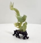 Escultura chinesa em jade natural, finamente talhada, representando uma ave adulta alimentando seu filhote, símbolo tradicional de proteção, prosperidade e continuidade familiar. A pedra apresenta tonalidade verde-clara suave com veios translúcidos característicos do jade, polimento acetinado e excelente aproveitamento das variações naturais da rocha para dar profundidade ao movimento das asas e definição ao pequeno filhote. A composição repousa sobre base tradicional em madeira entalhada à mão, com formas orgânicas que equilibram a verticalidade da peça. Dimensões: 22x16x7 centímetros. Apresenta pequenos desgastes e perdas em pontas mais delicadas, comuns em esculturas de pedra.
