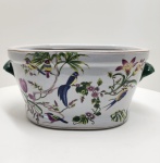 Faiança europeia decorativa. Peça em faiança esmaltada, ricamente decorada com motivos naturalistas de pássaros e flores tropicais entrelaçados por ramos sinuosos, em policromia viva sobre fundo branco. Datada entre 1900 e 1930, período marcado pela influência do estilo Chinoiserie e pelo renascimento das formas coloniais e naturalistas na arte decorativa europeia. A base apresenta relevo em forma de cruz (+), característico dos suportes utilizados durante a queima em fornos de alta temperatura nas manufaturas de faiança do final do século XIX e início do XX. O craquelé fino e uniforme da superfície do esmalte evidencia o envelhecimento natural da camada vítrea, atestando sua autenticidade e as técnicas tradicionais de produção. A ausência de marca de fábrica sugere uma peça destinada ao mercado de exportação ou a linhas populares de prestígio médio, prática comum em casas como Sarreguemines e Longchamp (França) ou Vista Alegre (Portugal) nas primeiras décadas do século XX. Dimensões: 27 x 44 centímetros.