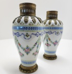 Elegante par de vasos franceses em porcelana fina da manufatura real de Sèvres, datados de 1771 conforme a marca de letra-data S pintada em azul sob o esmalte. As peças apresentam corpo ovóide delicadamente pintado à mão com guirlandas florais, laços azulados e festões de rosas em policromia suave, característicos da estética refinada de meados do século XVIII. Os vasos são montados em bronze dourado (ormolu) com aplicações de folhas estilizadas junto ao gargalo e bases circulares com frisos decorativos, reforçando o luxo típico das encomendas aristocráticas do período pré-Revolução. A paleta clara, a pincelada minuciosa e o equilíbrio das proporções evidenciam o trabalho de ateliês especializados da manufatura em sua fase áurea. Conjunto raro, harmonioso e absolutamente representativo do estilo Louis XVI. Dimensões: 20 x 10 centímetros cada.