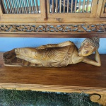 Escultura decorativa representando Buda reclinado, talhada em madeira maciça, com modelado suave e detalhes bem definidos nas vestes e no cabelo em pontos estilizados. A figura repousa sobre o próprio bloco de madeira, com o corpo alongado e a cabeça apoiada na mão direita, posição tradicionalmente associada ao Parinirvana (momento da passagem para o Nirvana final).A superfície apresenta acabamento natural em tom mel, com sombreados que realçam o entalhe e a movimentação do tecido. Dimensões: 25x82x20 centímetros.