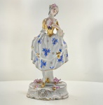 Figura feminina em porcelana Vista Alegre, Portugal, meados do século XX. Figura de dama oitocentista, em porcelana portuguesa moldada e pintada à mão, representando jovem segurando leque. Traje decorado com flores azuis e douradas, corselete em ouro brilhante, e base rococó com pequenas flores aplicadas em relevo. Marca em verde sob a base: V.A. Portugal (Vista Alegre). Dimensões:26 x 11 x 10 centímetros. Estado de conservação: apresenta reparo visível no pescoço, pequenas perdas nas flores aplicadas e leve desgaste do dourado, compatíveis com a idade.