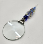 Lupa artística com cabo em vidro Murano, peça contemporânea em execução manual, apresentando cabo alongado em vidro soprado com aplicações de murrine multicoloridas e aventurina, terminando em esferóide azul profundo unido a ferragem decorada em metal prateado trabalhado; lente circular de amplo diâmetro, transparente e de boa graduação, formando conjunto elegante que combina utilidade e arte em um objeto refinado de caráter decorativo e colecionável. Dimensões: 20x6 centímetros.