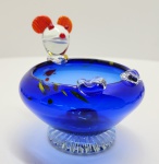Centro decorativo em vidro artístico soprado, possivelmente Murano, Itália, meados do século XX. Peça em vidro azul cobalto de corpo globular assentado sobre base moldada em relevo radial, decorada com inclusões multicoloridas internas. Na borda, três aplicações em vidro claro modelado manualmente e figura lúdica de palhaço igualmente em vidro, com cabeça translúcida e detalhes policromos em vermelho e amarelo, típica da produção de estúdios muraneses voltados ao estilo figurativo pós-guerra. Trabalho de execução cuidadosa, com bom equilíbrio entre forma, cor e fantasia estética. Dimensões: 13x13x13 centímetros.