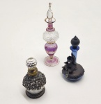 Conjunto de três delicados frascos antigos de perfume, cada um com características únicas de época e manufatura. O frasco em vidro roxo e transparente apresenta corpo torneado com aplicações douradas e cúpula rica em detalhes lapidados, acompanhado de um aplicador igualmente trabalhado à mão. O frasco azul possui base em vidro soprado com montura metálica ornamentada e figura feminina em relevo, revelando produção artesanal de inspiração europeia do início do século XX. O frasco leitoso, por sua vez, exibe corpo decorado com uma complexa armação metálica cravejada de pequenas pedras e tampa facetada, refletindo o estilo exuberante dos frascos de toucador vitorianos. Dimensões: 14x4, 10x5, 8x4 centímetros.