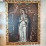 Rara e expressiva obra sacra do Século XV, representando a tradicional iconografia da Virgen Coronada, inserida no universo devocional da pintura hispano-cristã tardo-medieval. A composição apresenta a Virgem em posição central, frontal, com as mãos unidas em gesto de oração, envolta por amplo manto azul profundo. Seu rosto longo, delicado e de feições suavemente idealizadas é característico da estética pré-renascentista ibérica.Acima, dois anjos sustentam a coroa imperial, um motivo recorrente na arte medieval tardia que reforça a condição da Virgem como Rainha dos Céus. Nos flancos da composição aparecem reservas circulares com símbolos marianos construções, astros e elementos naturais típicos do repertório emblemático utilizado nas ladainhas e alegorias religiosas do período.Na parte inferior, figura a representação de um dragão estilizado, símbolo medieval da vitória do bem sobre o mal, frequentemente associado à iconografia da Imaculada ou da Virgen de la Expectación.A obra conserva uma pátina antiga marcante, com craquelure fina e escurecimento natural das camadas pictóricas, compatíveis com pinturas de origem medieval. A moldura entalhada e dourada, embora posterior, dialoga com o caráter ornamental da peça. Dimensões: