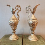 Par de jarras em metal dourado, ricamente trabalhadas em estilo Rococó, com corpo em formato ovóide decorado por baixos-relevos figurativos, representando cenas campestres, putti e elementos vegetalistas. Bicos alongados com figura de querubim em relevo e alças em volutas ornamentais. Bases largas, com frisos florais e acabamento em douração. Dimensões:50 x 21 x 15 centímetros.
