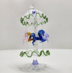 Carrossel em vidro artístico Murano Delicada escultura em vidro artístico de Murano, representando um carrossel com cavalos coloridos, executada com notável precisão artesanal. A estrutura translúcida é adornada por bordas onduladas em verde, sustentando delicadas figuras de cavalos em tons de âmbar, lilás e azul, modeladas à mão com transparência e movimento fluido. A composição expressa a leveza e o encanto do vidro soprado veneziano, evocando o universo lúdico e nostálgico dos antigos carrosséis. Altura: 15 x 7 centímetros.