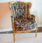 Poltrona finamente trabalhada em estilo Luís XVI, com estrutura em madeira entalhada e acabamento em folha de ouro, preservando o brilho e a riqueza decorativa típicos do neoclassicismo francês. O encosto apresenta formato envolvente, com laterais levemente abertas e moldura contornada por frisos, rosetas e entalhes de folhas. O assento é largo e confortável, revestido em brocado de padrão damasco, em tom bege com desenho em preto, combinando textura, elegância e presença decorativa. Os braços são delimitados por entalhes florais e volutas, acompanhando o desenho do encosto. A peça repousa sobre quatro pés canelados, adornados com motivos vegetalistas, finalizando em pequenos pés torneados elementos característicos do período Luís XVI.