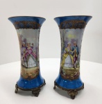 Par de vasos em porcelana SÈVRES, ambos marcados na base, decorados com sena galante pintada à mão e fundo clássico bleu de Sèvres. As reservas ovais apresentam sena galante executada com paleta suave e acabamento minucioso, contornada por rica douração aplicada a pincel. O corpo dos vasos exibe esmalte azul profundo com ornamentação dourada em volutas e arabescos característicos da manufatura. As peças repousam sobre bases em bronze trabalhado, com pés em volutas e acabamento condizente com as montagens francesas tradicionais. Interior das bocas com pintura dourada e motivos florais. Vasos marcados SÈVRES, conjunto de grande elegância e forte presença decorativa. Dimensões: 28 x 14 centímetros. ( um dos vasos está colado).