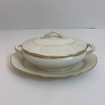 Conjunto composto por sopeira com tampa e travessa oval, produzido em porcelana portuguesa da manufatura VA Portugal. A peça apresenta acabamento esmaltado em tom marfim com frisos dourados, além de duas alças laterais e pegador superior na tampa, características típicas de serviços de mesa clássicos europeus do século XX.