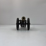 Miniatura de canhão decorativo, composta por estrutura em madeira com ferragens e rodas em metal, e tubo em bronze com acabamento envelhecido.Peça rica em detalhes, apresentando elementos simulados de funcionamento, como mecanismo de elevação e base com parafusos, remetendo a modelos históricos de artilharia.