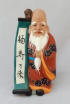CERÂMICA, uma (1) escultura policromada, pintada a mão, representando o Deus FUKUROKUJU, segundo o Xintoísmo, Mitologia e Folclore Japonês, Deus da longevidade e sabedoria, representado por uma longa barba branca trazendo na mão um cetro ou bastão (saku) sagrado onde esta pendurado um pergaminho (makimono) contendo as escritas da sabedoria mundial, considerado Deus da ecologia, as vezes acompanhado por um veado, tartaruga ou uma garça tipo grou (tsuru), outras vezes de um pêssego, ter sua estatueta traz popularidade e garante longevidade, integrante do Shichi Fukujin (os 7 Deuses da Boa Sorte, Fortuna e Felicidade), medindo 14 cm altura.