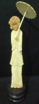 Escultura cor marfim oriental de dama segurando sombrinha com base de madeira enegrecida. Sec. XX. Alt.  34cm