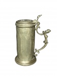 ESTANHO, um (1) tankard confeccionado em estanho, corpo em caneluras, alça em arabesco decorado por figura relevada, pega da tampa representando concha e tampa cinzelada por brasão heraldico, leves mossas na base, medindo 24x10cm.