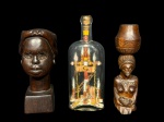 ESCULTURAS, lote com três (3) esculturas, sendo: A) duas (2) esculturas em madeira entalhada, representando figuras humanas de inspiração africana, uma em formato de busto feminino sobre base e outra de figura feminina com recipiente apoiado sobre a cabeça, medindo 17cm e 18cm e B) uma (1) garrafa decorativa em vidro, contendo composição interna artesanal com elementos geométricos e simbólicos, fechada com rolha, medindo 20cm.