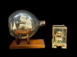 ESCULTURAS, lote com duas (2) esculturas, sendo: A) 1 navio em garrafa, miniatura artesanal de embarcação à vela, acondicionada em garrafa de vidro, com base em madeira, apresentando velas em tecido e casco em madeira, medindo 20x18cm e B) 1 vitrine oriental, contendo escultura decorativa representando pagode/templo asiático, confeccionada em materiais naturais (possivelmente madeira, fibras e elementos esculpidos), protegida por caixa de vidro com base ornamentada, medindo 10,5x6x6cm (falha na madeira).