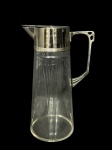 CRISTAL e METAL PRATEADO, uma (1) jarra estilo art noveau confeccionada em cristal translucido com lapidação de frisos horizontais, palmas e elementos geométricos em baixo relevo com gargalo e alça em metal espessurado a prata, medindo 27cm de altura.
