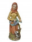 ESCULTURA, uma (1) grande escultura confeccionada em biscuit representando camponesa idosa, segurando avental com frutas (uvas e maçãs), acompanhada por cachorro aos pés, ricamente modelada e policromada, medindo 39cm de altura.