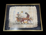 QUADRO, uma (1) reprodução em papiro, representando cena de inspiração egípcia, com figuras em carruagem, personagens a pé e elementos simbólicos acompanhados de escrita hieroglífica, moldura em metal dourado com proteção em vidro, medindo 42,5x55cm. Vidro quebrado.