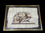 QUADRO, uma (1) reprodução em papiro, representando cena de inspiração egípcia, com figura em carruagem puxada por cavalos, acompanhada de elementos simbólicos e escrita hieroglífica, moldura em metal dourado com proteção em vidro, medindo 42,5x55cm.