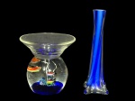MURANOS, lote com duas (2) peças confeccionadas em vidro de Murano, sendo: A) 1 castiçal translucido com parte inferior representando aquário com peixes policromados sobre fundo azul, medindo 13x14cm e B) 1 vaso solifleur confeccionado em vidro de Murano na tonalidade azul com moldagem torcida, medindo 20cm de altura.