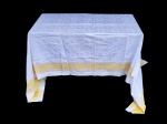 CAMA, MESA & BANHO, uma (1) toalha de mesa em tecido branco, apresentando padronagem em relevo por todo o campo e faixa decorativa na tonalidade amarela aplicada na parte inferior com motivos geométricos, leves manchas de guardado, medindo 312x292cm.