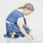 Bing & Grondahl, Copenhague, Dinamarca(Fundada em 1853). Grupo escultórico em porcelana esmaltada, representando figura infantil feminina ajoelhada, trajando vestido de mangas bufantes, com cintura marcada por faixa, em atitude de alimentar felino doméstico disposto à frente, junto a comedouro circular integrado à composição. Modelagem naturalista, com atenção às dobras do vestuário e à postura inclinada do tronco. Policromia executada sob esmalte, predominando as tonalidades azul, branco e tons de pele, com cabelos moldados e levemente sombreados. Base integrada à escultura, de contorno irregular, simulando apoio natural do solo. Peça moldada em corpo único. Apresenta ínfimo bicado na orelha direita do felino. Marca da manufatura, e numeração 1745, localizadas no verso. Medida: Alt.: 14 x Comp.: 15 x Larg.: 11 cm.